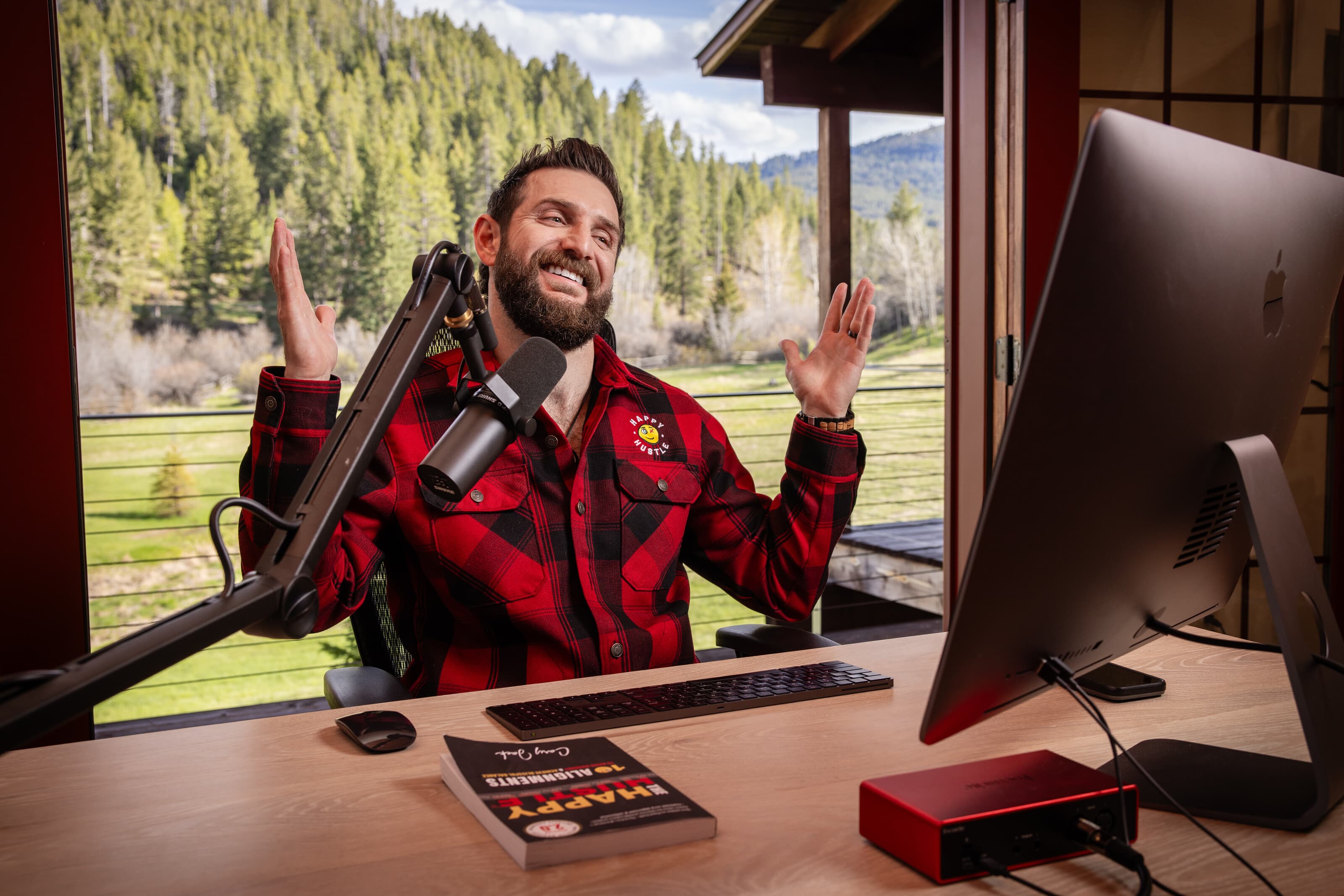 Cary Jack recording The Happy Hustle Podcast at his studio desk, Montana forest behind him